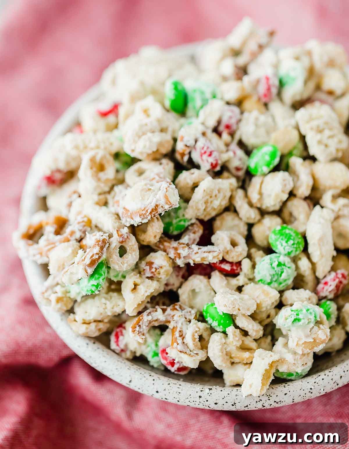 A black and white speckled bowl filled with reindeer chow, set on a vibrant red linen background, showcasing its festive appeal.