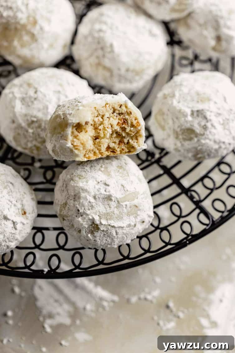 Freshly baked Snowball Cookies cooling on a wire rack, with one cookie bitten in half revealing its nutty interior.