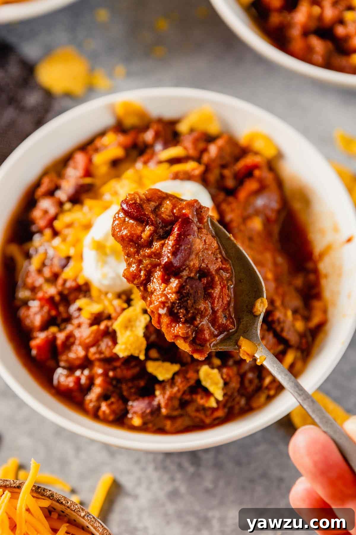 Close up photo of spoonful of chili held over a whole bowl of chili with sour cream and corn chips visible.