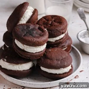 A square photo of a white plate with whoopie pies piled on top with a small glass of milk in the back.