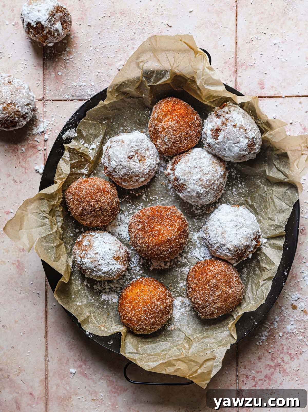 Overhead photo of fried zeppole coated in powdered sugar and cinnamon sugar in parchment paper in a dark pan.