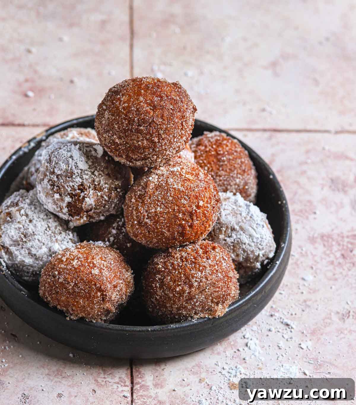 Fried zeppole tossed in powdered sugar and cinnamon sugar stacked in a low black bowl.
