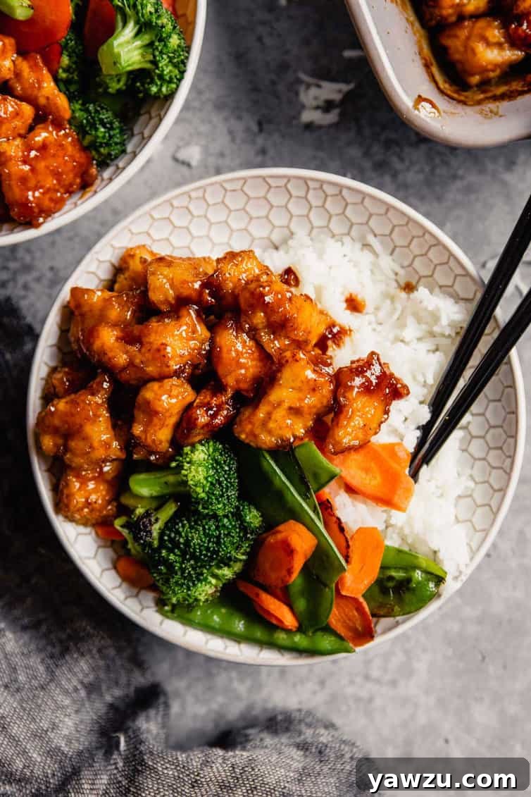 A white bowl filled with steamed rice, colorful stir-fry vegetables, and glistening baked sweet and sour chicken, ready to be enjoyed with chopsticks.