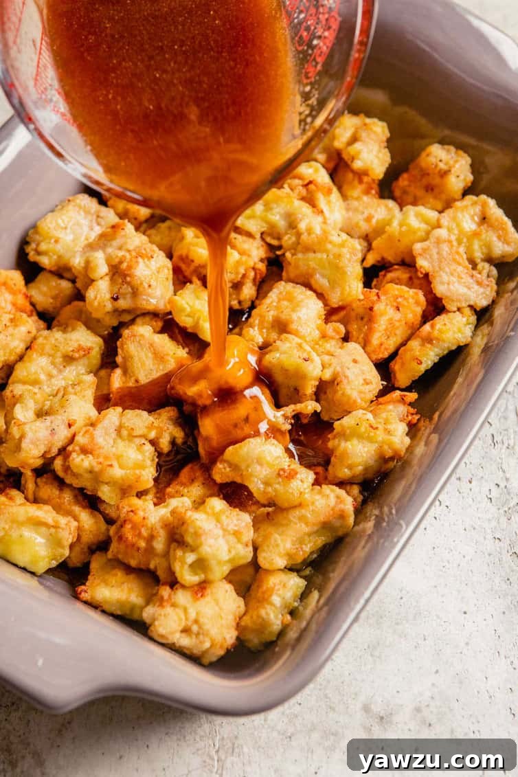 A 9x13-inch baking dish filled with golden-brown fried chicken pieces, as a generous stream of homemade sweet and sour sauce is poured over them, preparing for the final baking step.