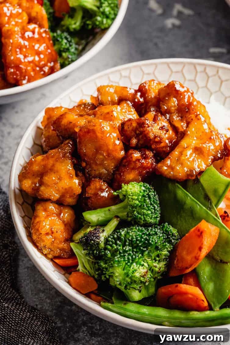 A beautifully arranged white bowl featuring sweet and sour chicken and vibrant stir-fry vegetables, positioned in front of another serving bowl on a stylish gray counter.