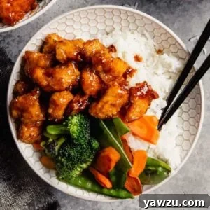 A white bowl showcasing baked sweet and sour chicken, accompanied by steamed broccoli, rice, and chopsticks, indicating a complete and delicious meal.