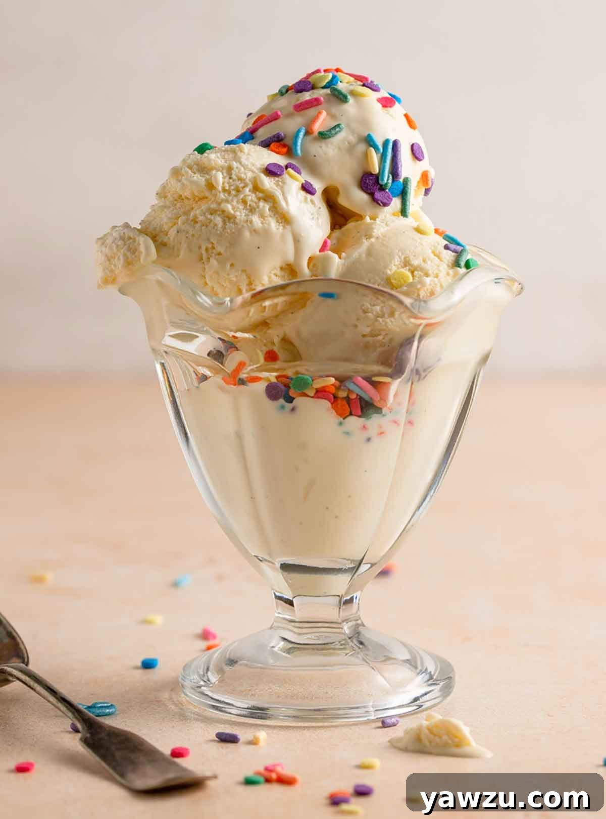 Three scoops of vanilla ice cream in a glass parfait dish with colorful sprinkles on top.
