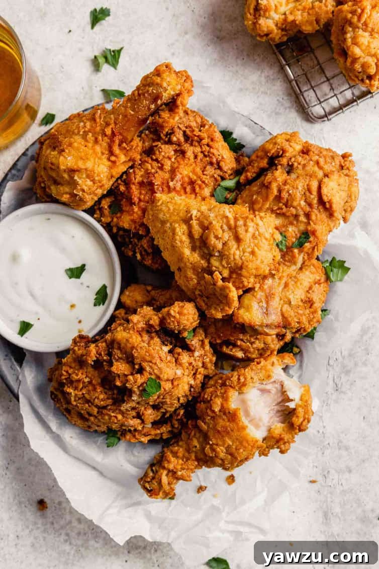 A fried drumstick on the right with a bite taken out at the bottom of a plate of buttermilk fried chicken with a small cup of ranch to the left.
