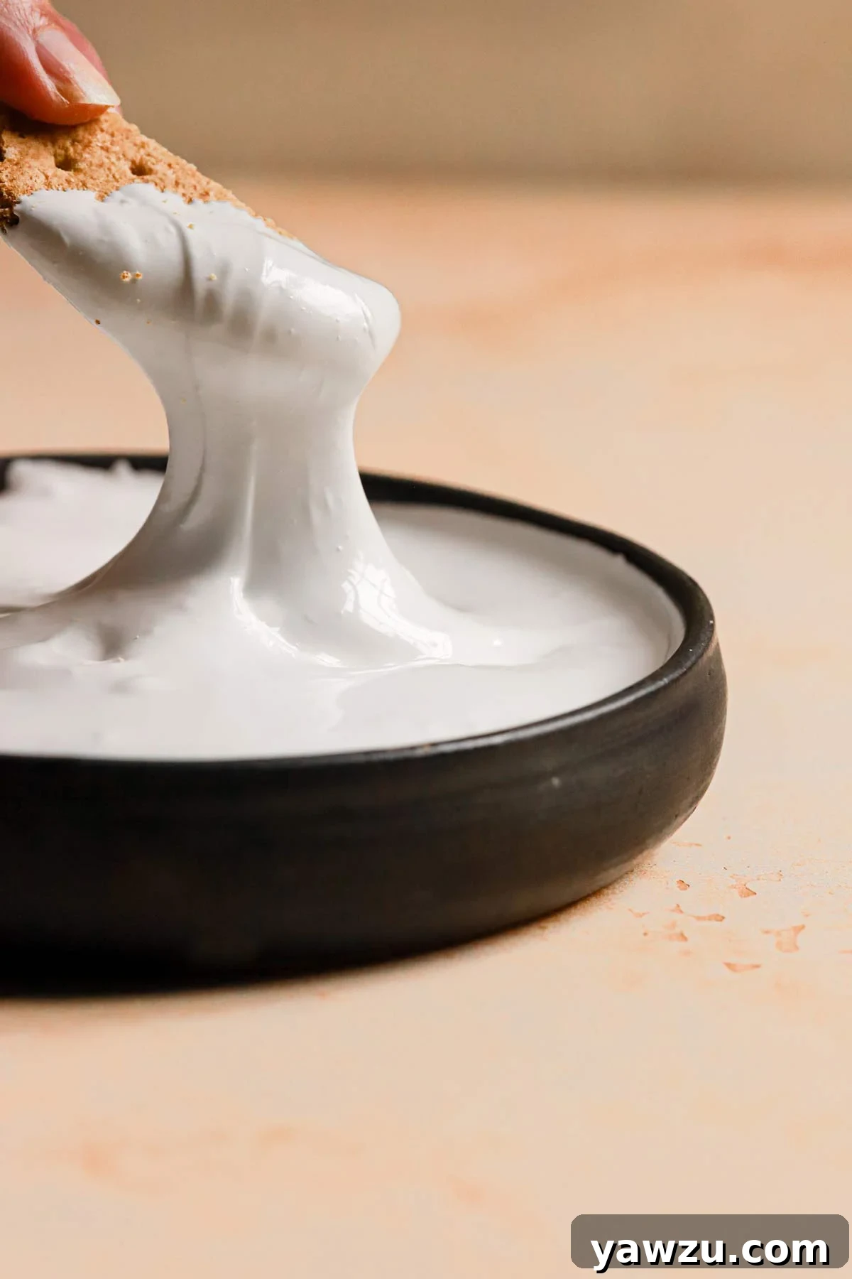 A black bowl on a peach counter with a graham cracker dipping into the marshmallow cream.