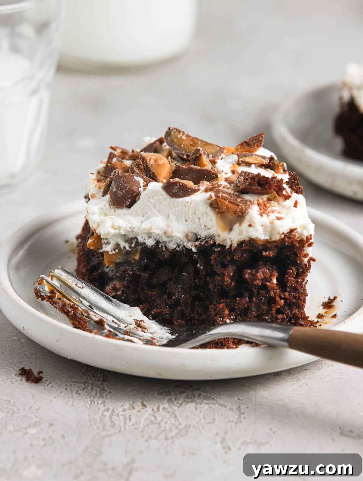 A single slice of better than sex cake on a rimmed white plate with a fork, showcasing its layers of chocolate cake, caramel, whipped topping, and crushed Heath bars.
