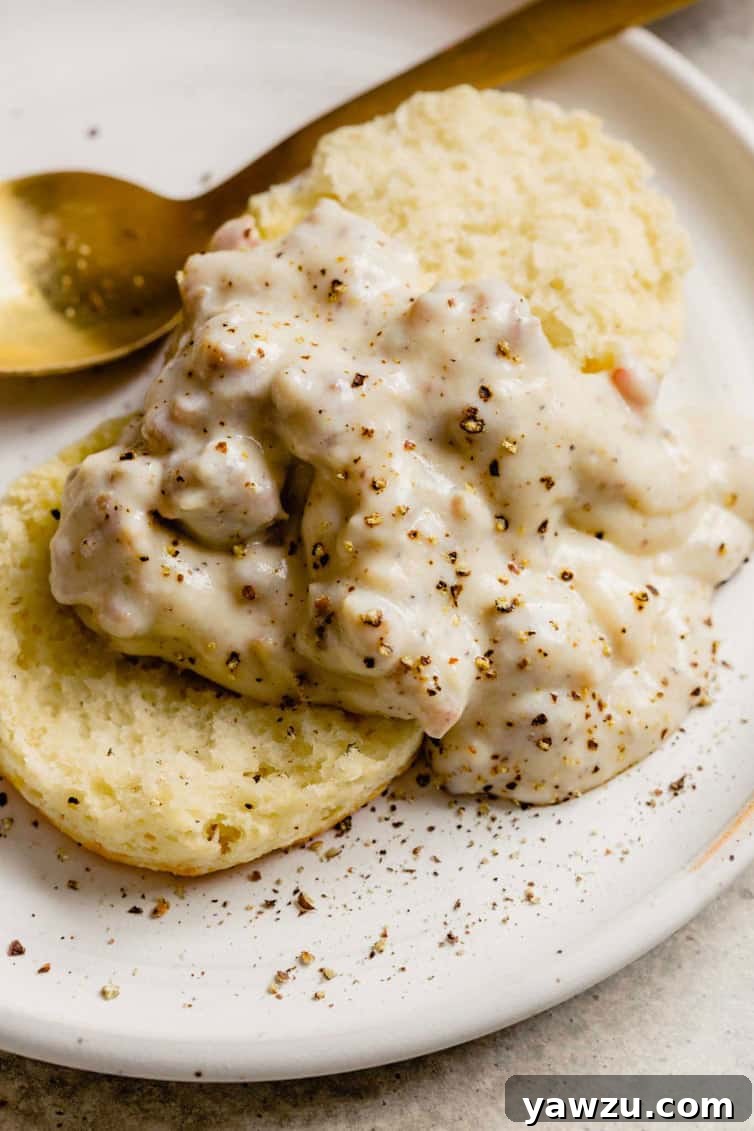 A top down picture of biscuits and gravy on a white plate with a gold spoon in the back. The gravy is thick and chunky with sausage pieces.