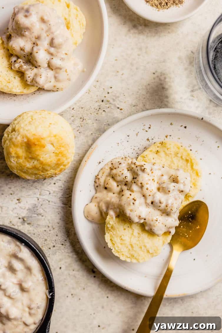 A plate of biscuits and gravy with a gold spoon on the right, a biscuit to the left of the plate, and another plate with biscuits and gravy in the top left. The focus is on the creamy texture of the gravy.