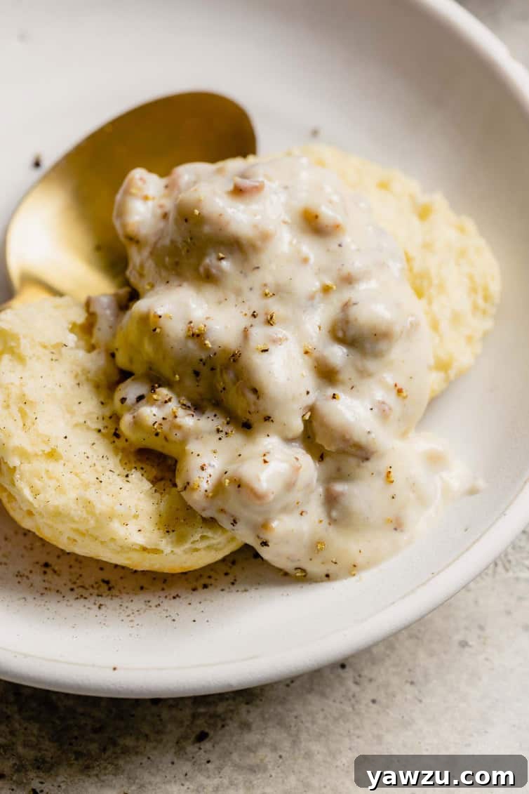 A close up shot of sausage gravy on top of an open biscuit on a white plate, highlighting the creamy texture and chunks of sausage.