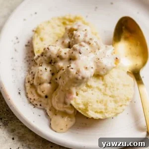 A biscuit topped with sausage gravy on a white plate with a gold spoon on the right.