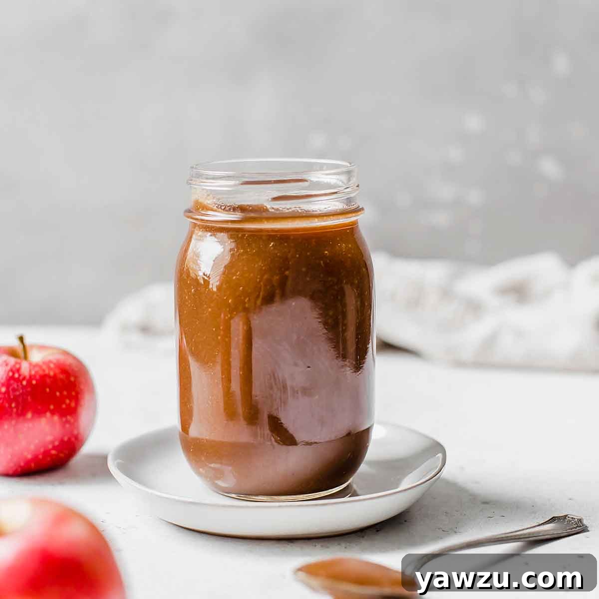 Jar of apple butter on a white saucer with a spoonful of apple butter in the foreground and two apples next to the jar.