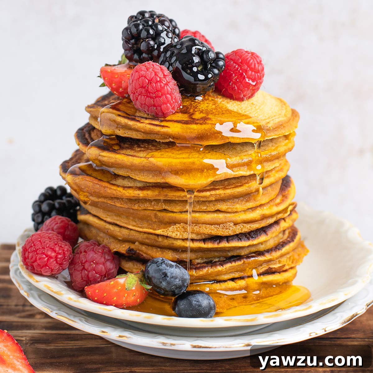 Stack of golden pumpkin pancakes on a white plate, generously drizzled with maple syrup, and garnished with fresh berries like blueberries and raspberries on top and around the base.