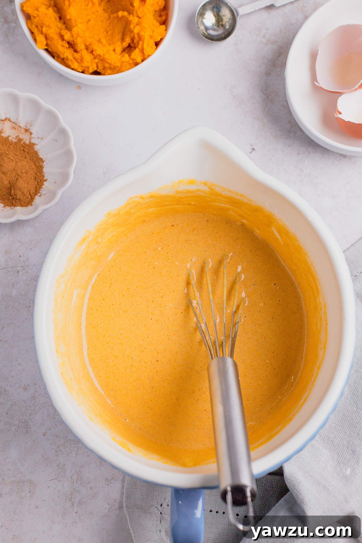 Close-up shot of pumpkin pancake batter in a large mixing bowl with a whisk. Around the bowl are smaller bowls holding pumpkin puree and various spices, prepped and ready for mixing.