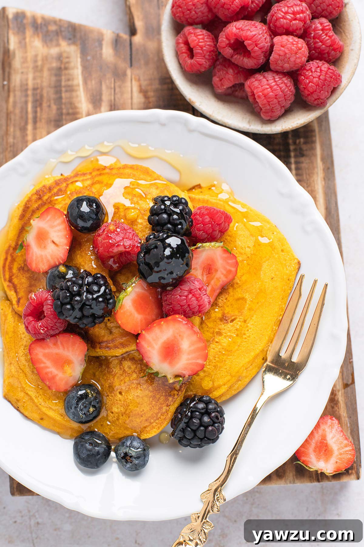 A perfectly styled plate of fluffy pumpkin pancakes, artfully arranged with a sprinkle of fresh berries on top and a generous, glistening drizzle of maple syrup flowing down the sides.