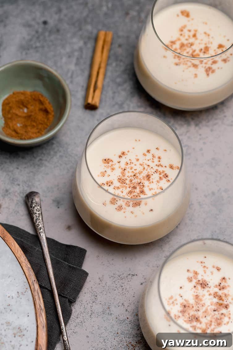 Three glasses of eggnog with a cinnamon stick and a bowl of grated nutmeg.