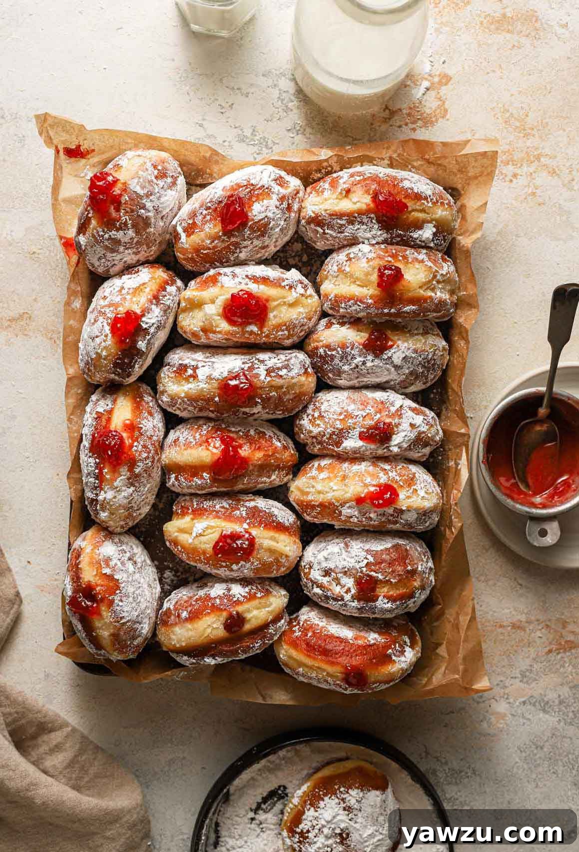 Overhead photo of paczki lined up on a parchment-lined baking sheet with a jar of milk, bowl of preserves, and powdered sugar surrounding it.
