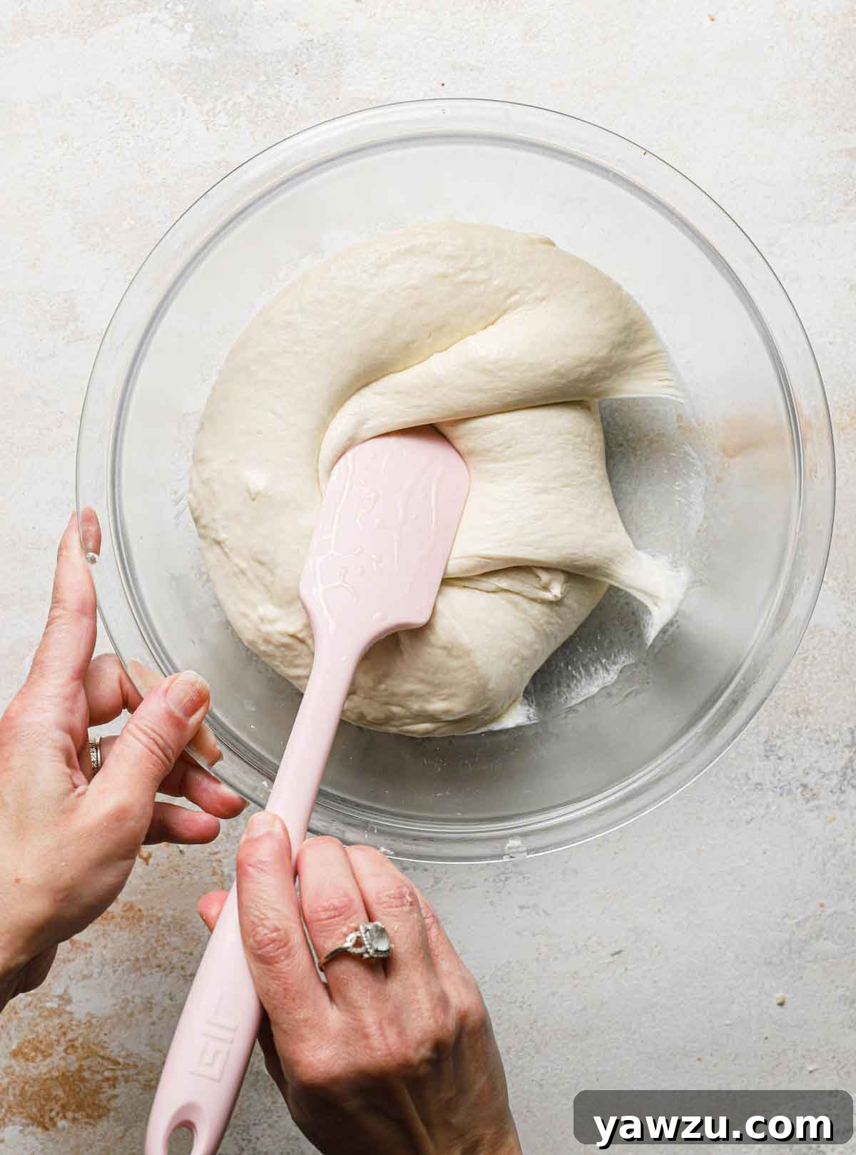 A rubber spatula being used to gently fold ciabatta dough over itself in a glass bowl, illustrating the folding technique.