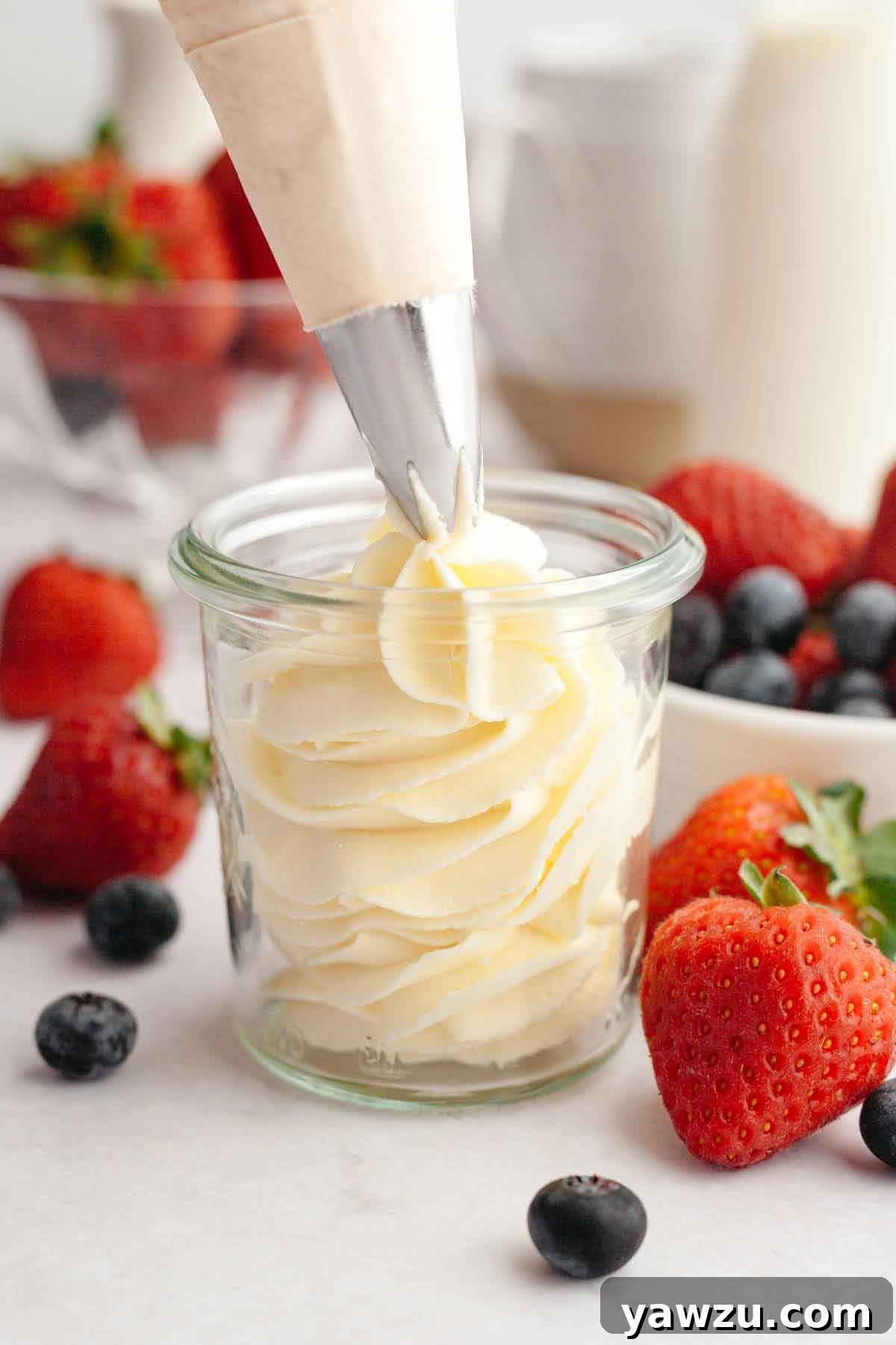 Homemade whipped cream being piped into a glass jar with strawberries and blueberries in the background.