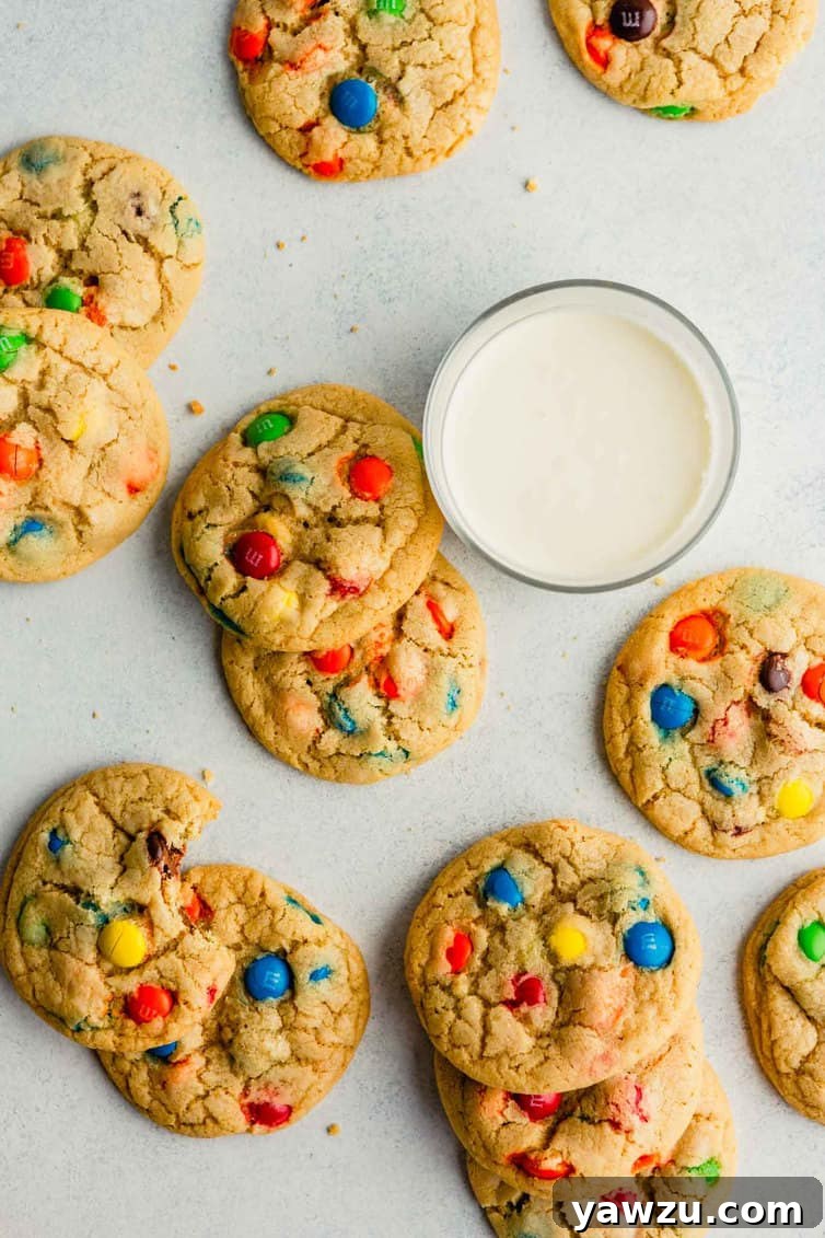 An overhead photo of freshly baked, colorful M&M cookies on a counter with a glass of milk, emphasizing their perfect chewiness.