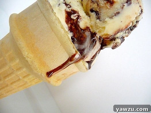 A scoop of homemade Tin Roof Ice Cream in a cone, with visible fudge swirls and chocolate-covered peanut chunks.