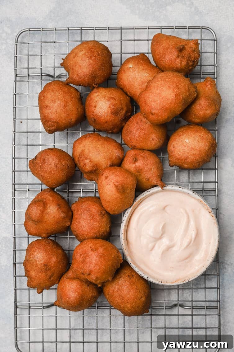 Freshly fried hush puppies cooling on a wire rack, with a bowl of dipping sauce in the background.