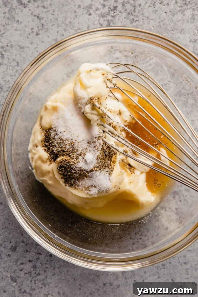 Glass bowl with a whisk on a grey counter with the coleslaw dressing before whisking.