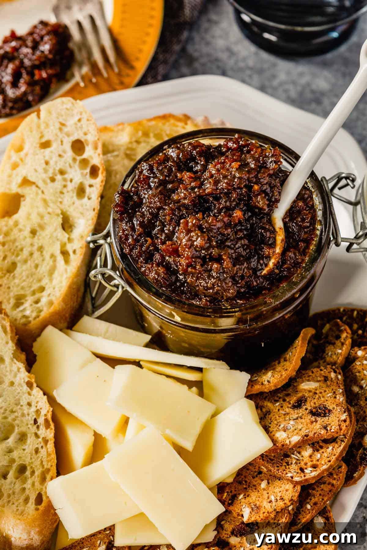 A glass jar of homemade bacon jam with a spoon on a white plate, flanked by cheese and crackers, ready to be served.