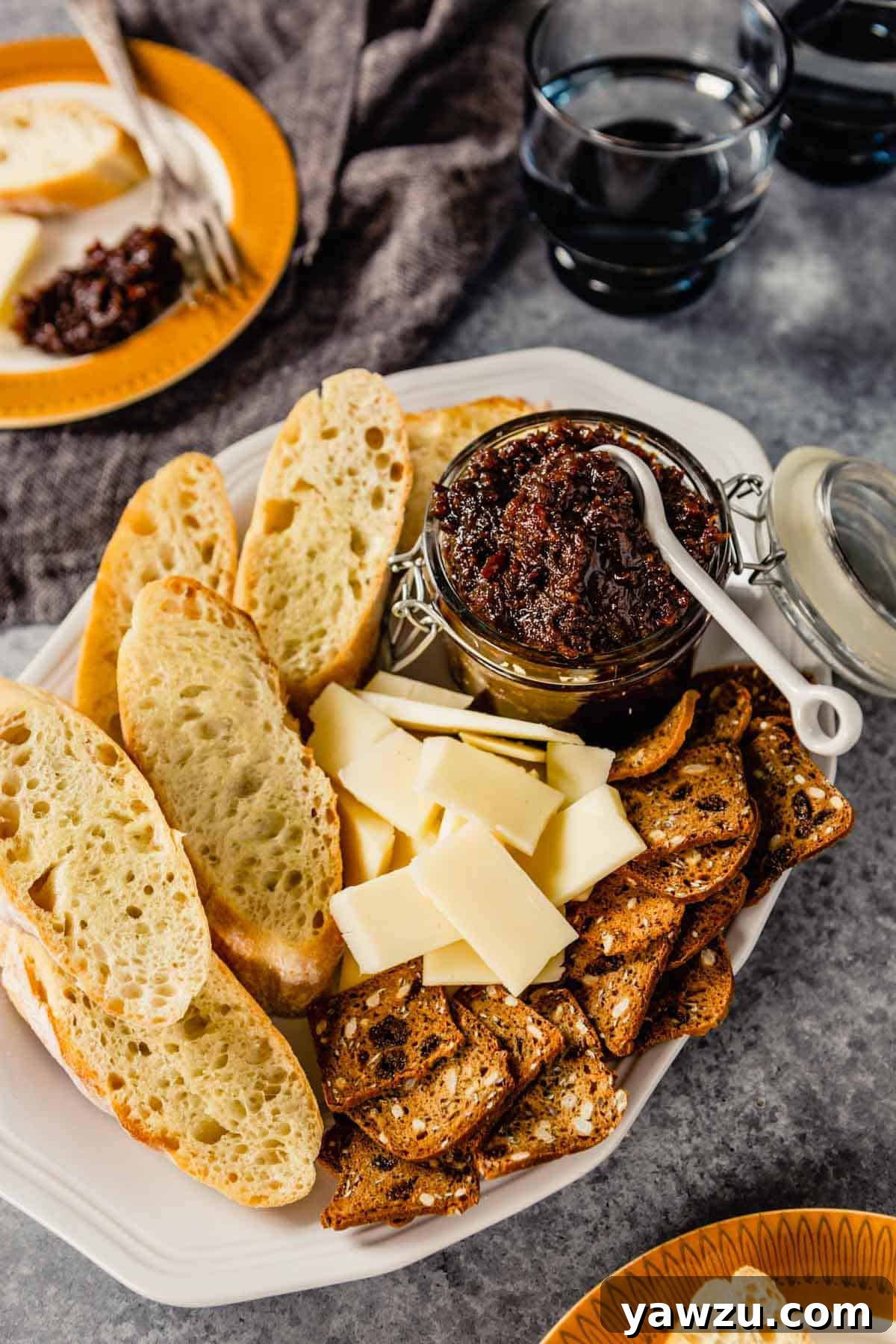 A festive platter featuring a jar of homemade bacon jam, surrounded by an assortment of cheeses, crackers, and slices of bread, perfect for entertaining.