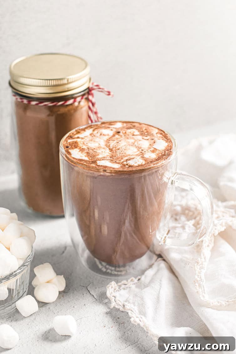 A beautifully styled cup of hot chocolate, generously topped with fluffy marshmallows, with a clear glass jar of the homemade hot chocolate mix blurred in the background, creating a cozy and inviting scene.
