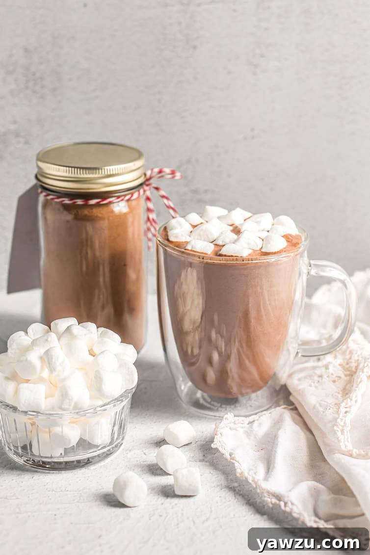 A charming close-up of a cozy cup of hot chocolate, lavishly topped with an abundance of mini marshmallows, while a jar of the homemade hot chocolate mix is gently blurred in the background, creating a warm, inviting, and comforting visual narrative.