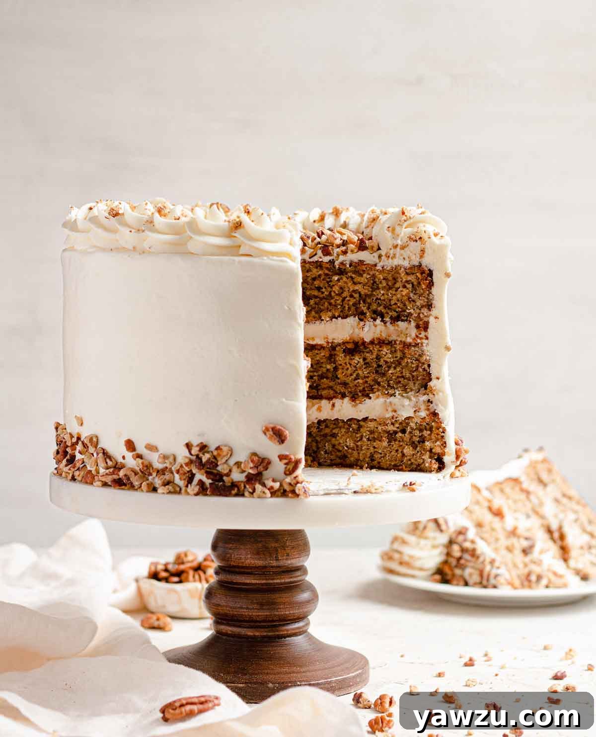 Side view of three-layer hummingbird cake with a slice removed, filling and frosted with cream cheese frosting and garnished with chopped pecans.