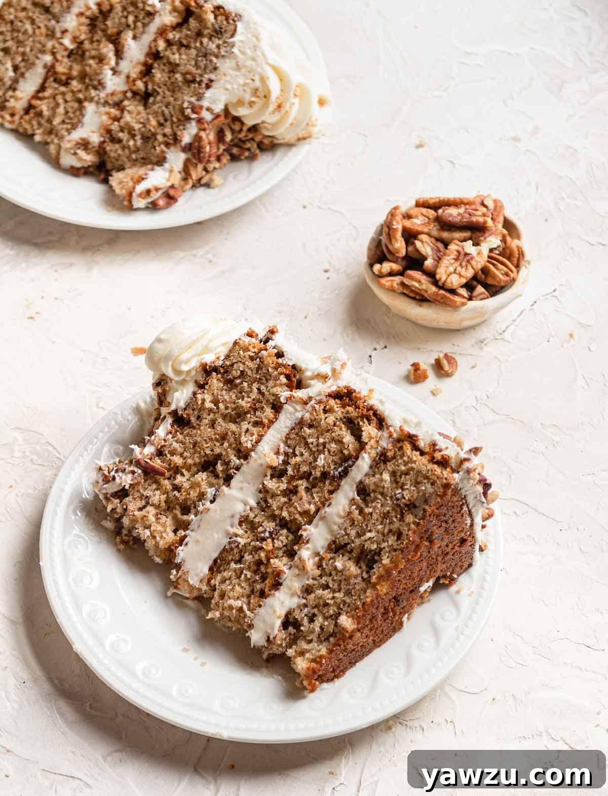 Slice of hummingbird cake on a white plate with a small bowl of pecans in the background.