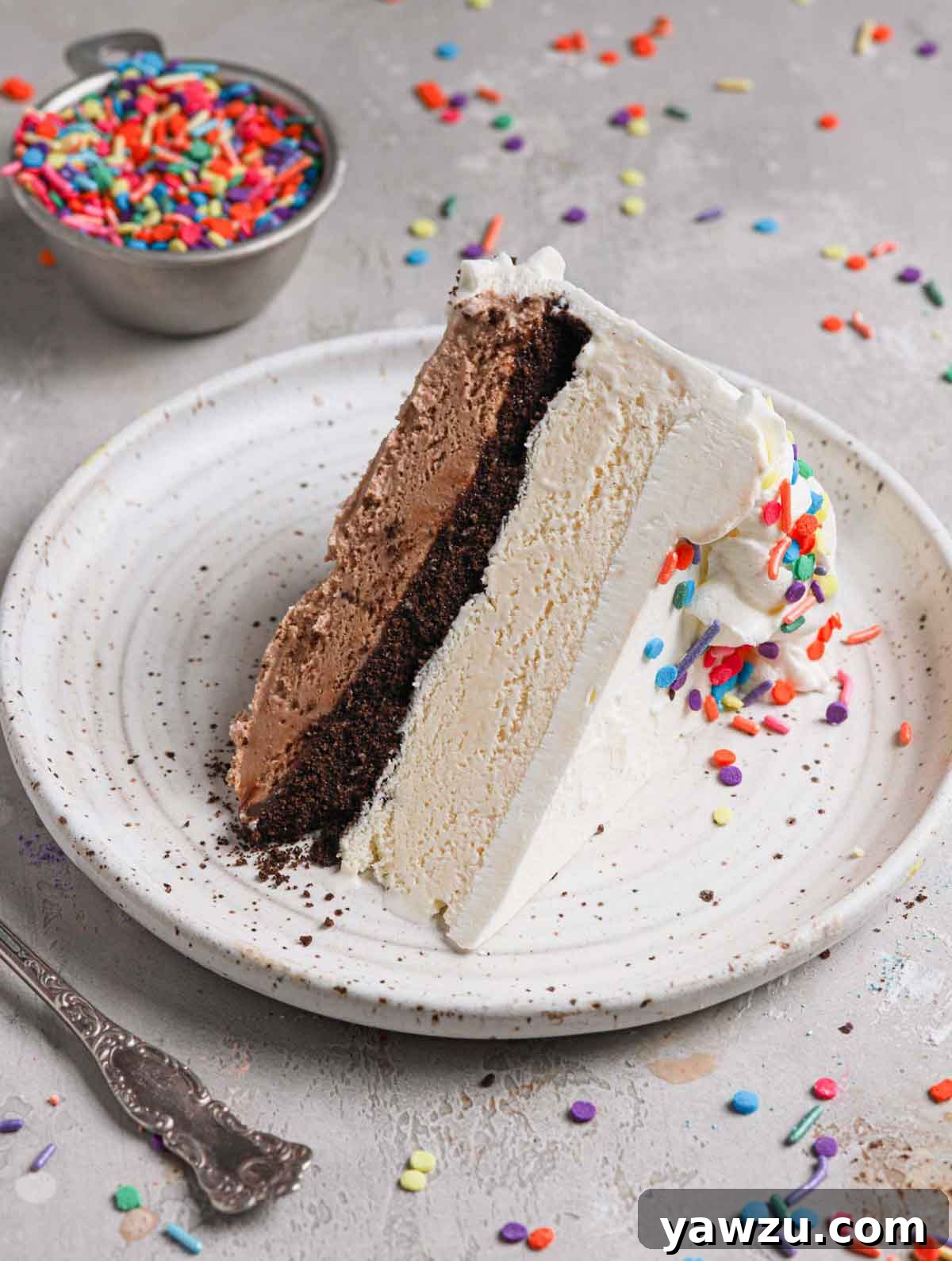 A perfectly sliced homemade ice cream cake revealing distinct layers of chocolate ice cream, vanilla ice cream, hot fudge, and crushed Oreos, served on a white plate with colorful sprinkles.