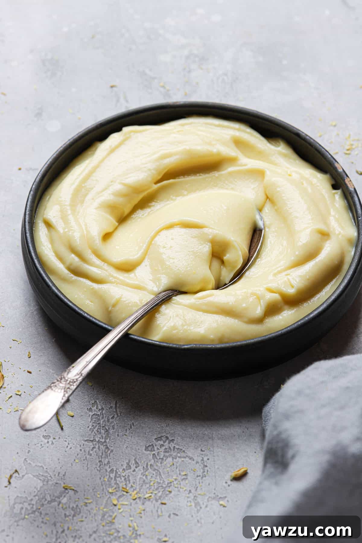 A black bowl of pastry cream with a spoon in the left side of the bowl on a grey counter with a light grey towel.