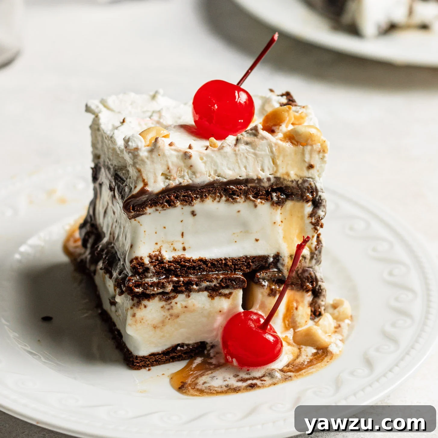 A perfectly square slice of ice cream sandwich cake on a white plate, garnished with a bright red maraschino cherry and chopped peanuts, showcasing its beautiful layers.