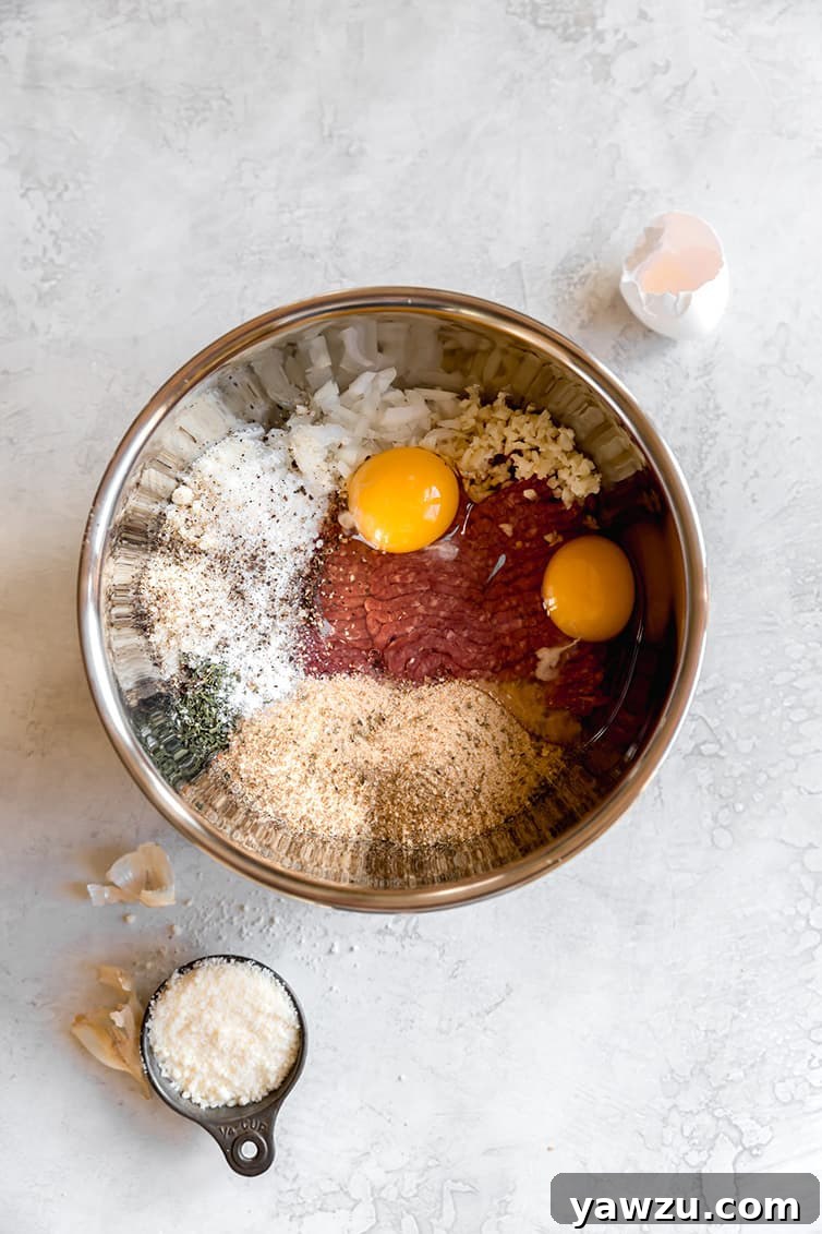 Various ingredients for an Italian meatball recipe, including ground meats, breadcrumbs, and cheese, in a large stainless steel mixing bowl.