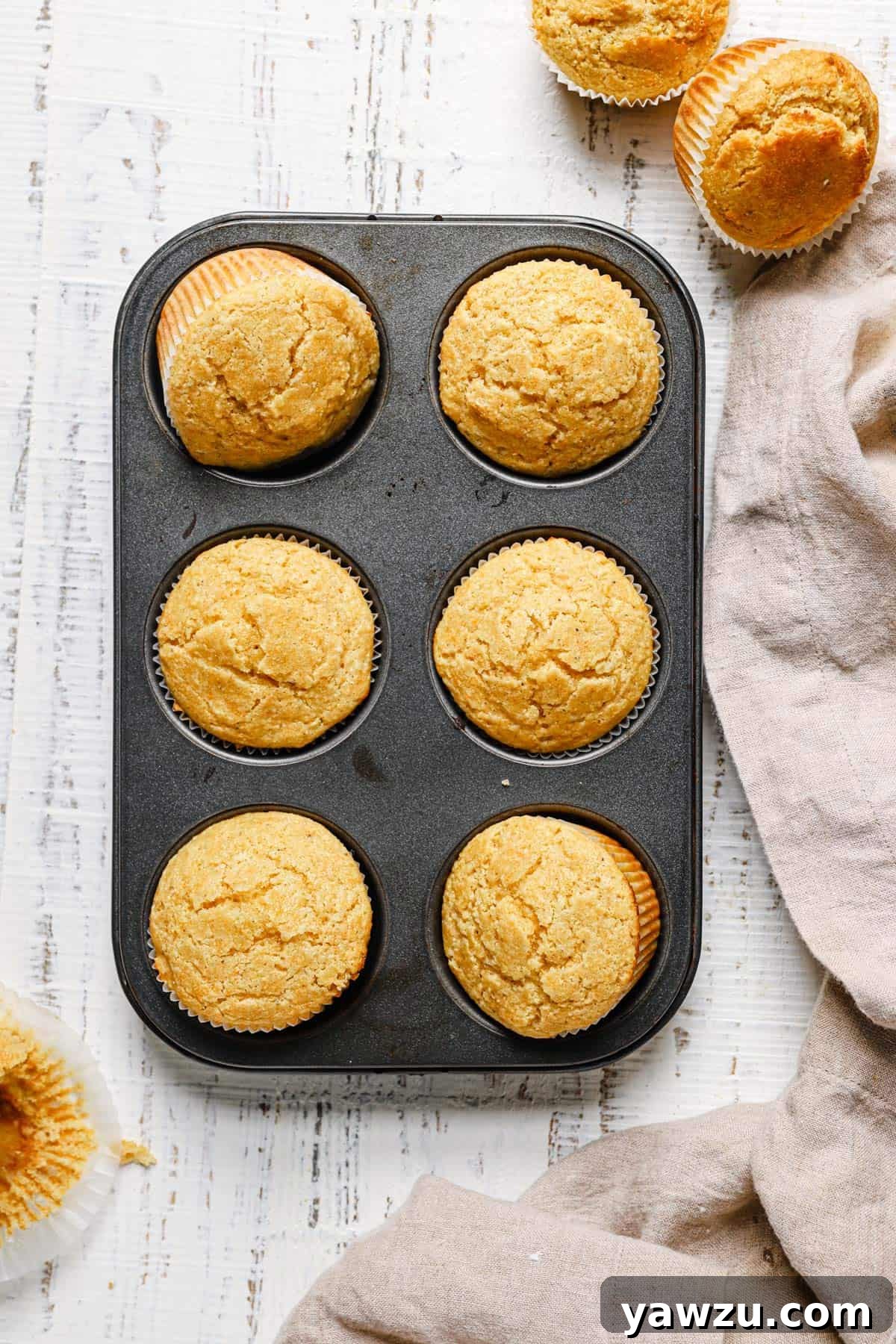 Top down photo of a muffin tin with 6 cornbread muffins on a white counter with two muffins in the top right and a towel in the bottom right.