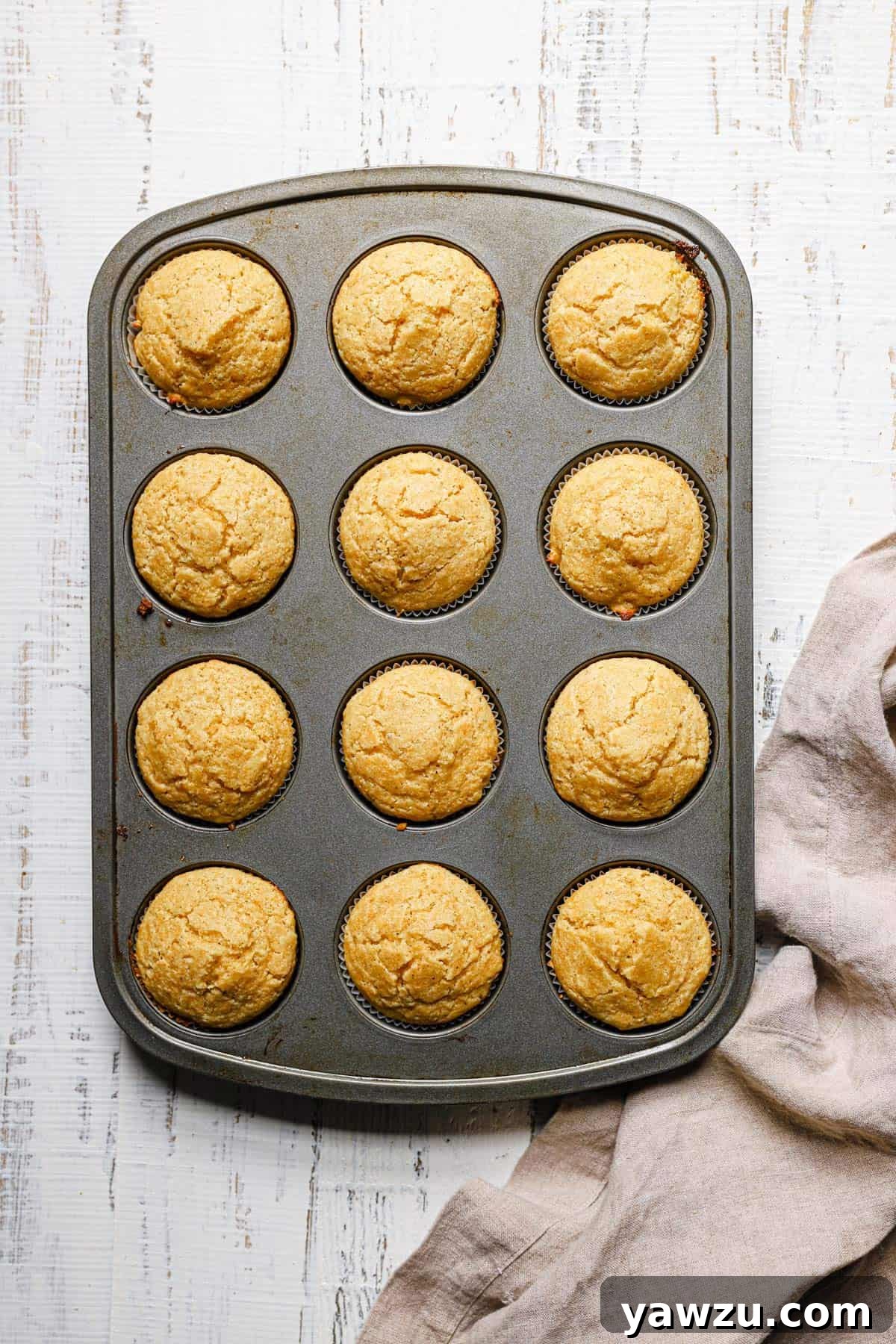 A silver muffin tin with cornbread muffins on a white counter with a light beige dish towel to the left.