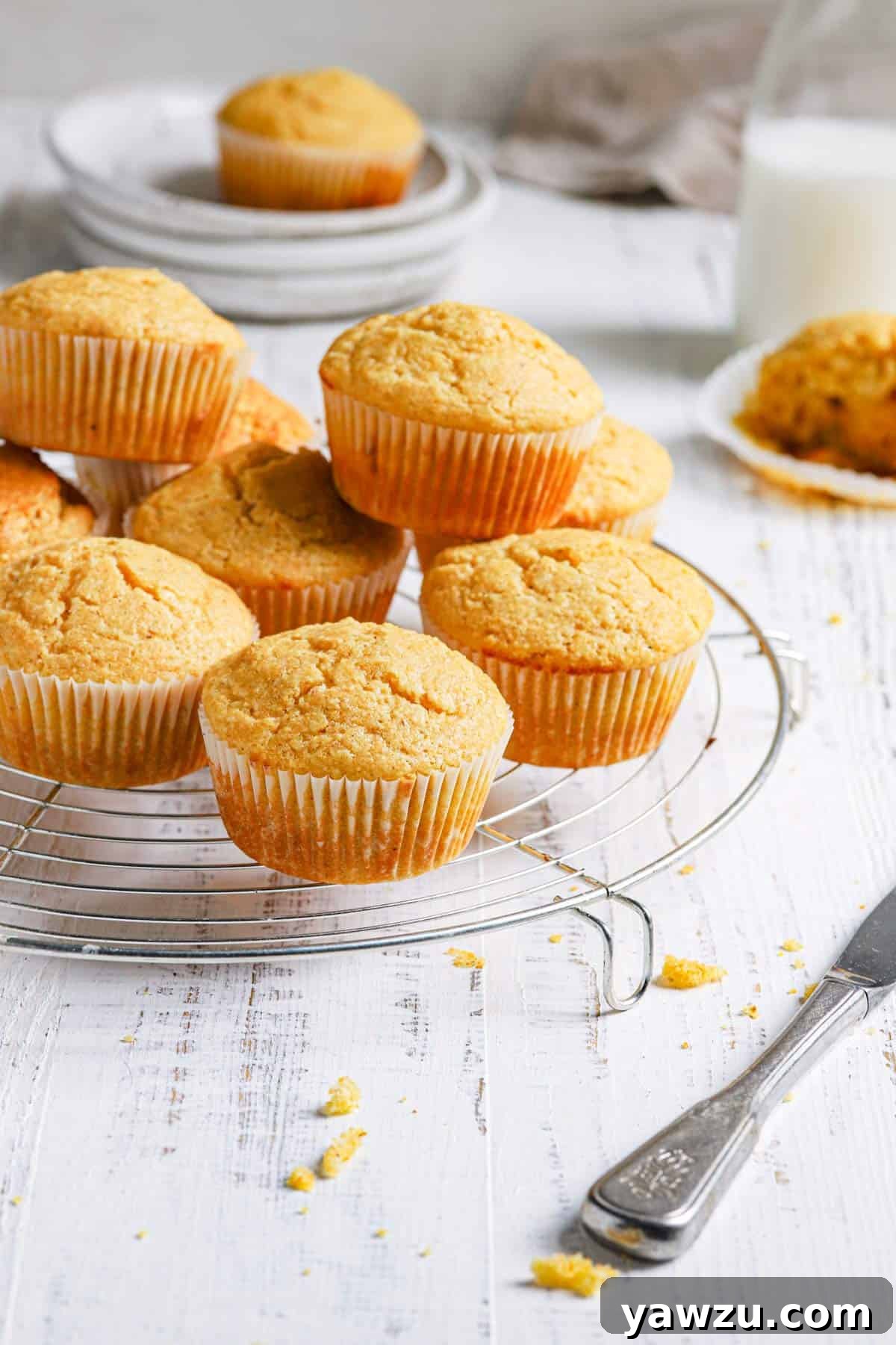 A wire cooling rack with cornbread muffins in a stack with a stack of plates in the back topped with a cornbread muffin and a milk jug to the right.