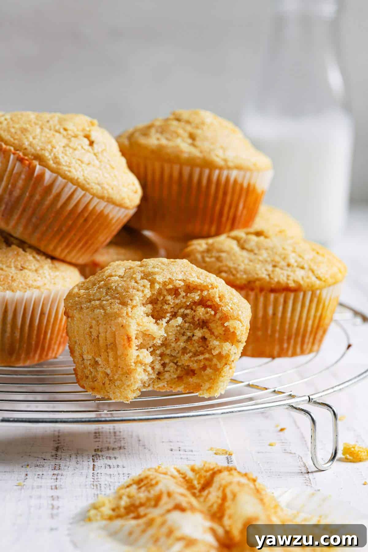 Fresh cornbread muffins on a wire rack with the front muffin missing a bite and a jug of milk in the back.