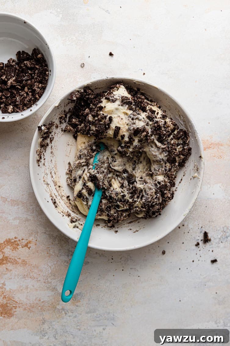 Close-up shot of creamy white cream cheese frosting, generously studded with dark, crushed Oreo cookies, in a mixing bowl.