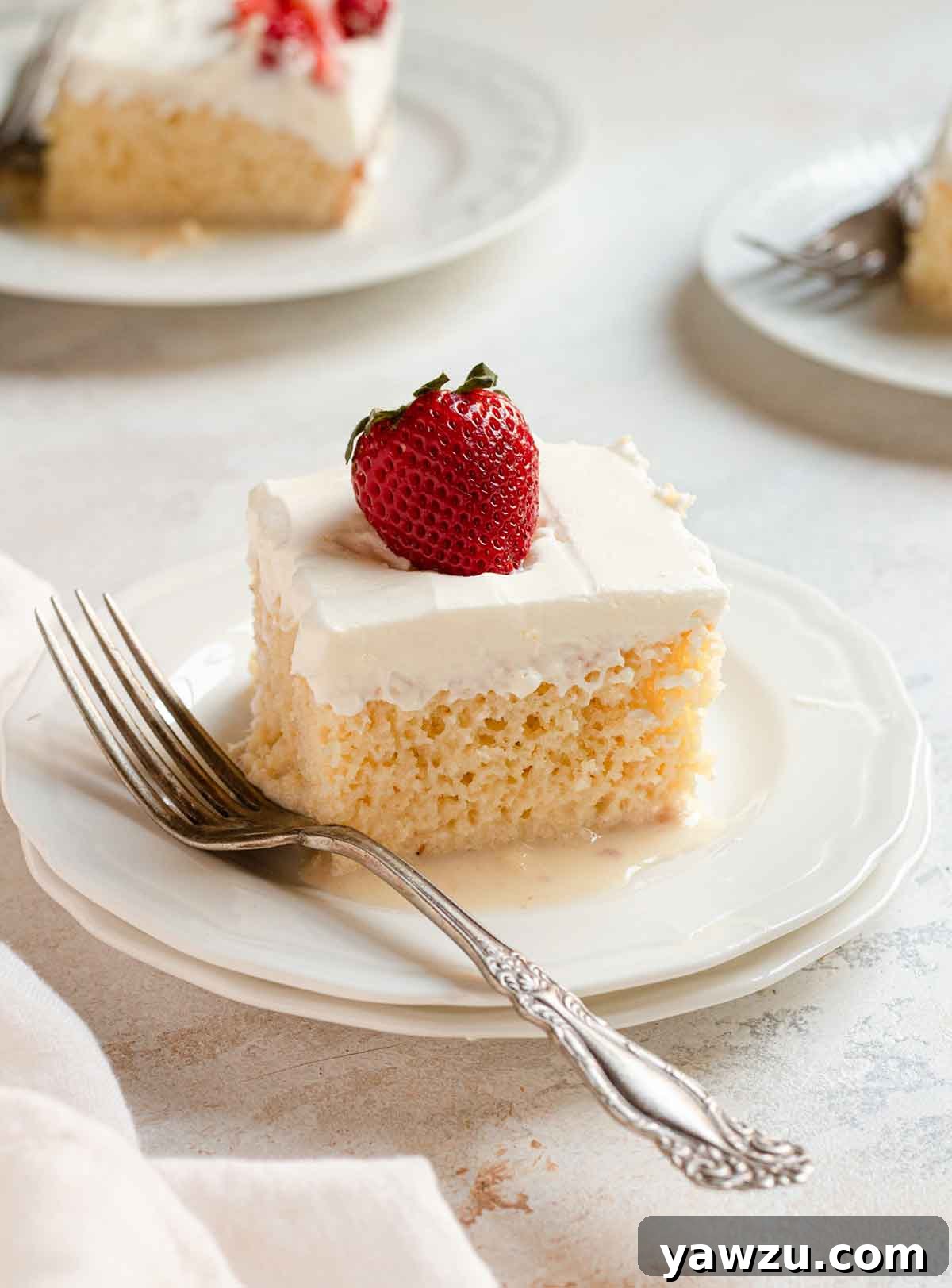 A piece of tres leches cake topped with a strawberry on a white plate with a fork on the side.