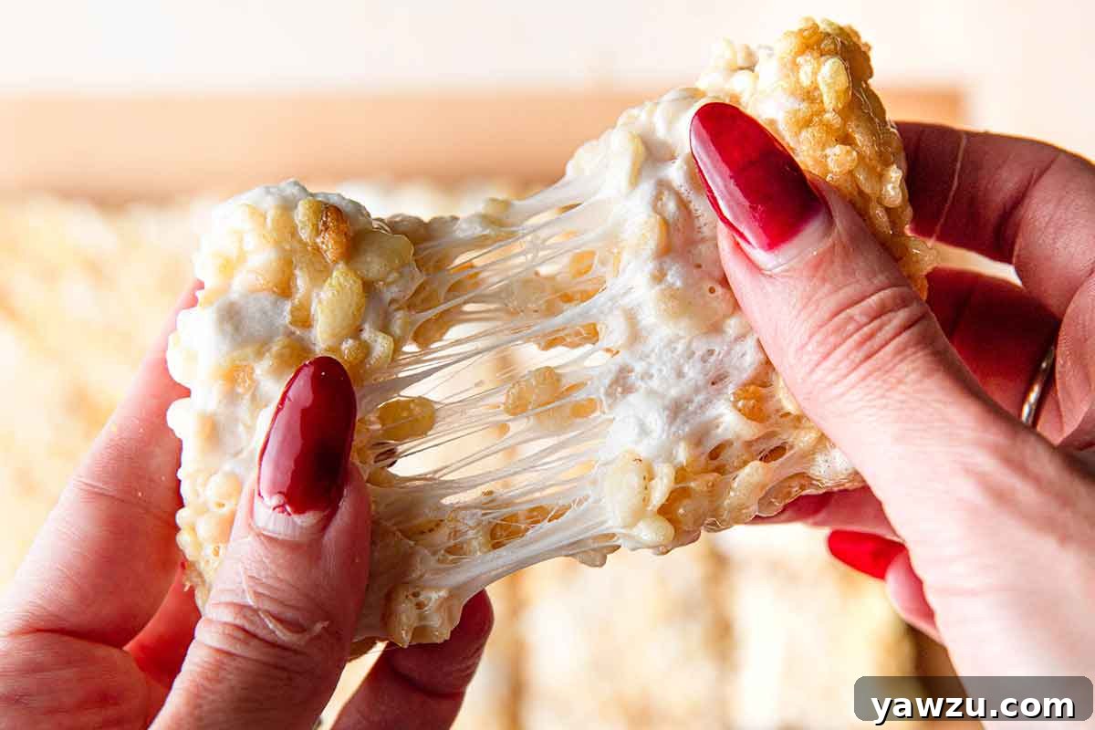 A perfectly gooey rice krispie treat being pulled apart, revealing luscious strings of melted marshmallow in the middle, emphasizing its chewy texture.
