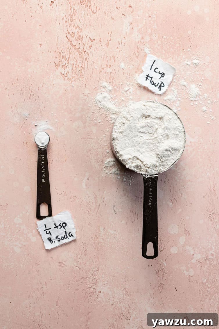 A measuring spoon with baking soda next to a measuring cup of flour.
