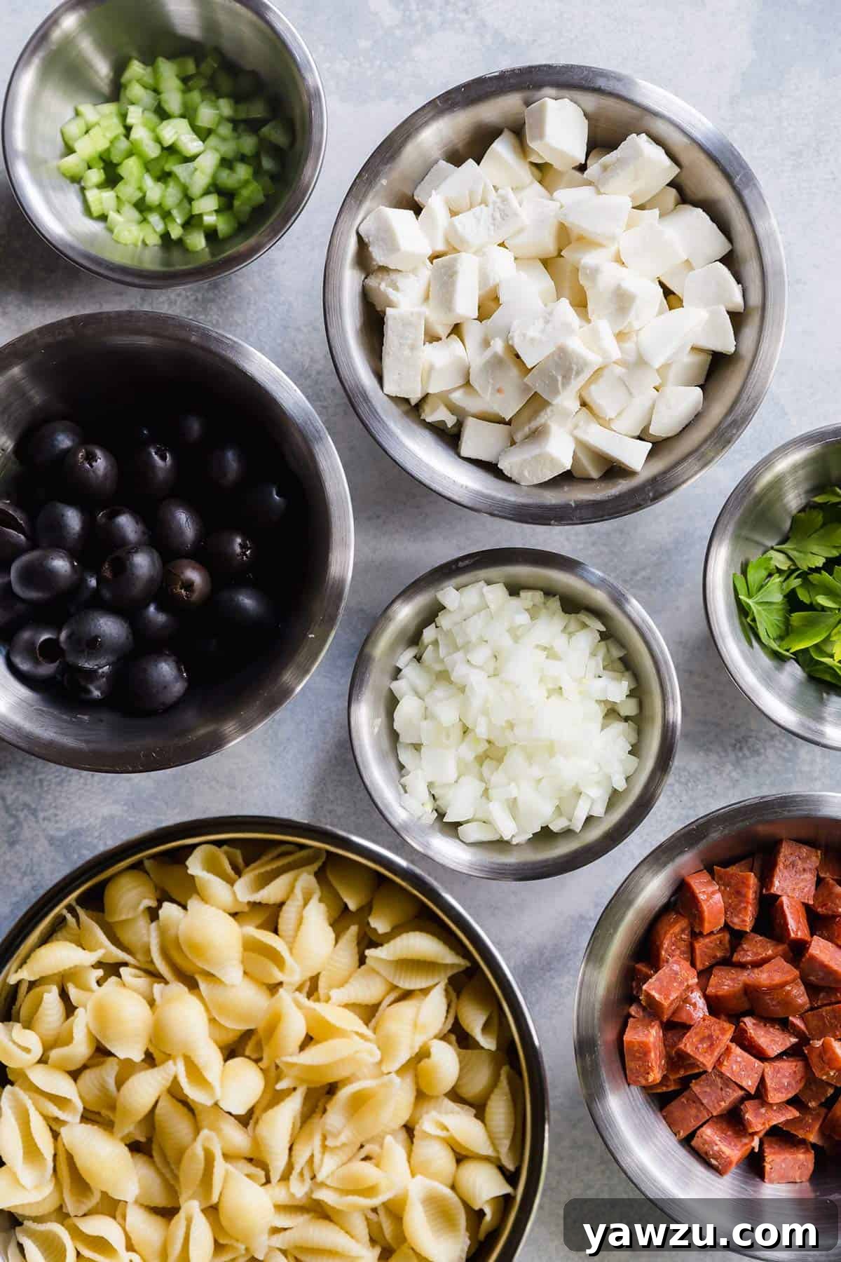 Fresh ingredients for Italian pasta salad, including cooked pasta, sliced pepperoni, cubed mozzarella, chopped onion, celery, olives, and fresh parsley, neatly prepped in bowls on a kitchen counter.