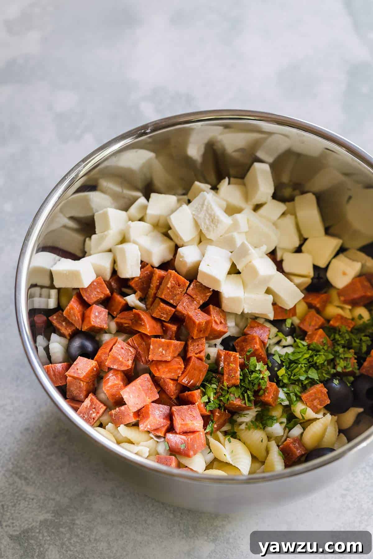 All the prepped ingredients for Italian pasta salad, including pasta, pepperoni, cheese, and vegetables, are gathered in a large mixing bowl, ready to be combined.